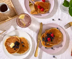 Plan en plongée de trois assiettes de pancakes garnis de petits fruits, disposées sur une table avec des ustensiles dorés, des p