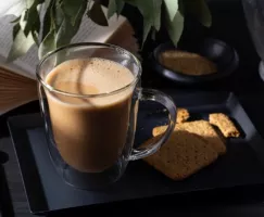 Tasse en verre remplie de kopi cham sur un plateau avec des biscuits, un livre et une plante