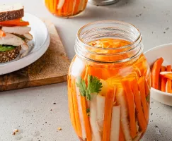 Un bocal de carottes et de radis daikon marinés avec un sandwich en arrière-plan.
