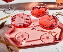 Trois cocktails pétillants à la grenade et à la rose dans des verres sur un plateau rose.