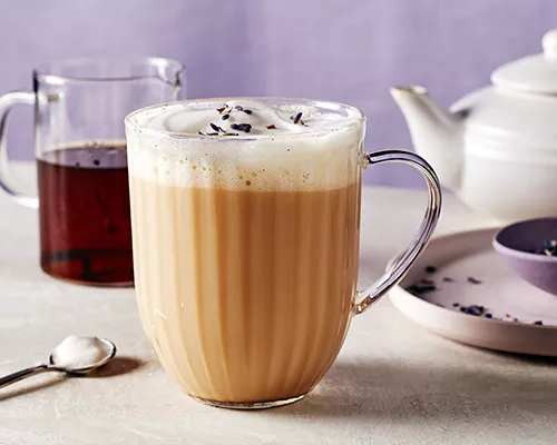 Un verre à latte London Fog avec mousse de lait, montré avec une carafe de sirop de vanille, une théière, une cuillère à mélanger et un bol pourpre de thé Earl Grey en vrac sur un plateau.
