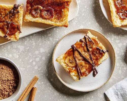 Quatre tartes renversées à la banane, deux dans des assiettes et deux sur un plateau, présentées avec un bol de sucre brun et des bâtonnets de cannelle.
