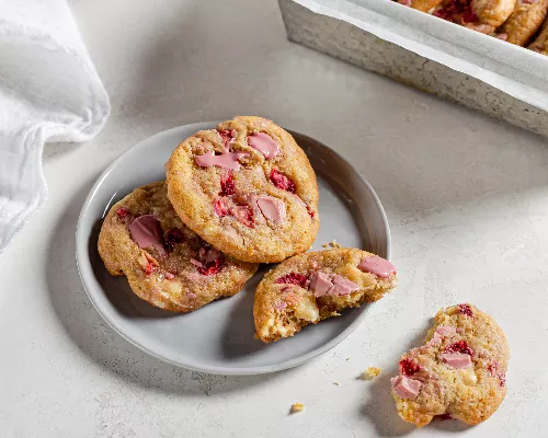 Trois cookies aux fraises et au chocolat rubis sur une assiette, dont un cassé en deux. 